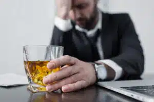 Man sitting at his desk in front of a laptop with a drink in his hand.