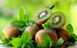 A pile of whole kiwis with one cut in half on top of the pile sitting on a table.