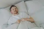 View from above of lonely upset young bearded man lying in bed with open eyes and can't sleep, feeling unhappy and tired, squeezes blanket with hands.