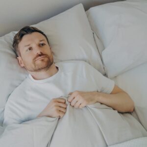 View from above of lonely upset young bearded man lying in bed with open eyes and can't sleep, feeling unhappy and tired, squeezes blanket with hands.